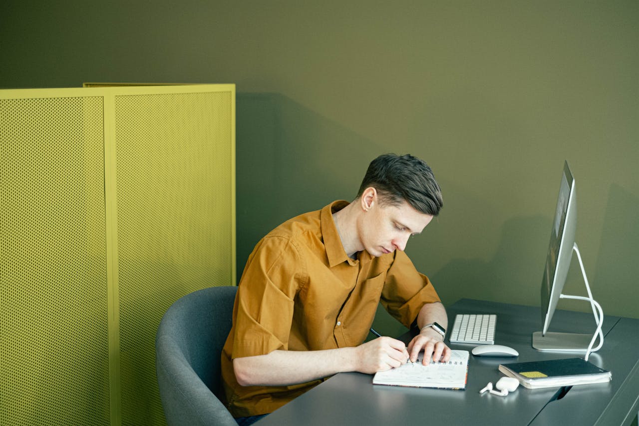 digital A man in a brown shirt sketching in a notebook at a tidy desk in a modern office setting.